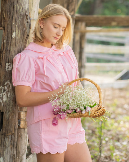 Woman wearing the Rosebud cotton pyjama set by Simply Provincial — soft pink button-up top with Peter Pan collar and delicate bows on the chest, paired with scalloped-edge shorts. Handmade from natural cotton and designed to capture the charm of slow, nostalgic mornings.