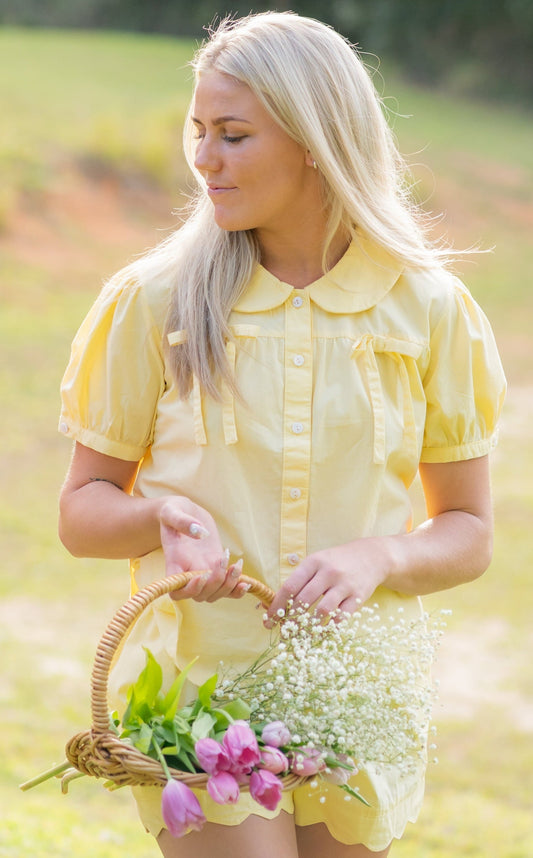 Woman wearing the Honeydrop cotton pyjama set by Simply Provincial — soft yellow button-up top with Peter Pan collar and delicate bows on the chest, paired with scalloped-edge shorts. Handmade from natural cotton and inspired by the warmth of slow, sunlit mornings.