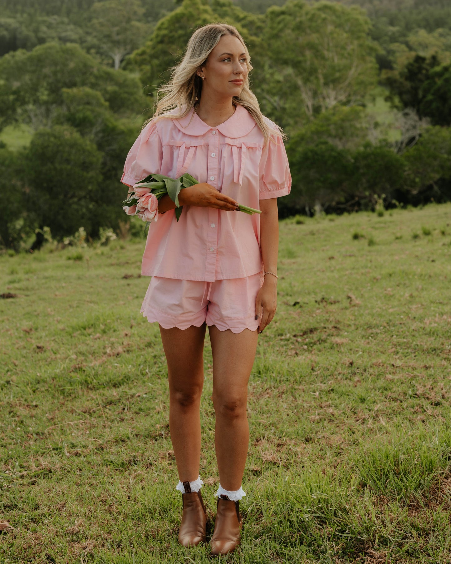 Woman wearing the Rosebud cotton pyjama set by Simply Provincial — soft pink button-up top with Peter Pan collar and delicate bows on the chest, paired with scalloped-edge shorts. Handmade from natural cotton and designed to capture the charm of slow, nostalgic mornings.