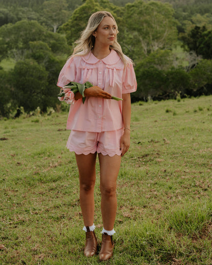 Woman wearing the Rosebud cotton pyjama set by Simply Provincial — soft pink button-up top with Peter Pan collar and delicate bows on the chest, paired with scalloped-edge shorts. Handmade from natural cotton and designed to capture the charm of slow, nostalgic mornings.