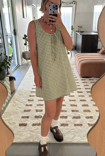 Person wearing a green gingham cotton dress taking a mirror selfie in a room with a patterned rug. The dress has an adjustable neckline which is feeding friendly for mums. 